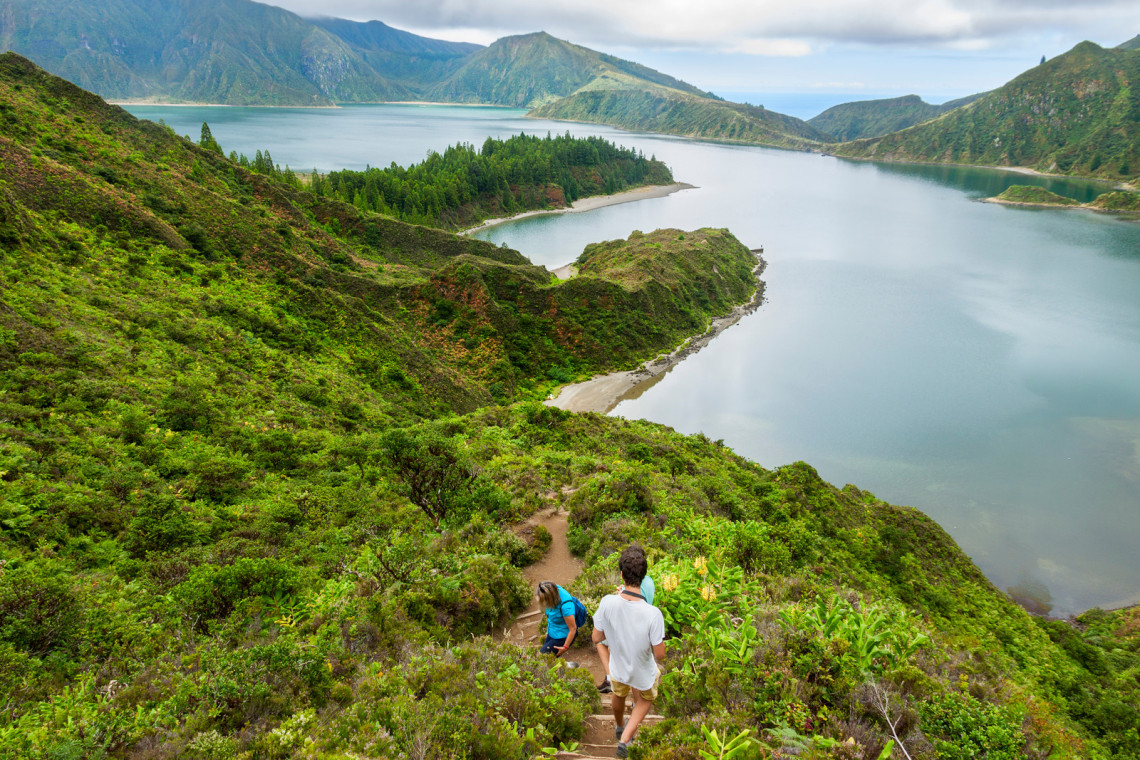 Hikers descending a narrow trail above Lagoa do Fogo, surrounded by lush vegetation with panoramic views across the volcanic crater lake.