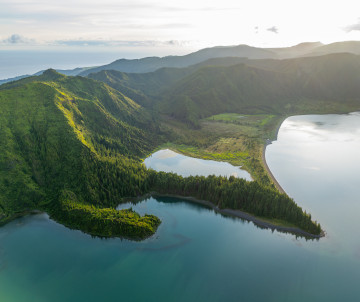 Aerial view of Lagoa do Fogo on São Miguel Island, showing blue crater waters, a forested peninsula and steep green volcanic ridges.