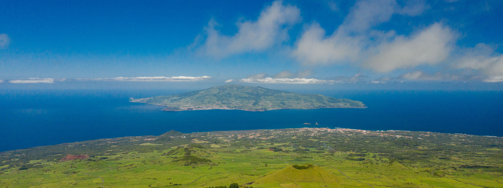 Elevated view from the slopes of Mount Pico across the green landscape of Pico Island and the Atlantic channel toward neighboring Faial Island.