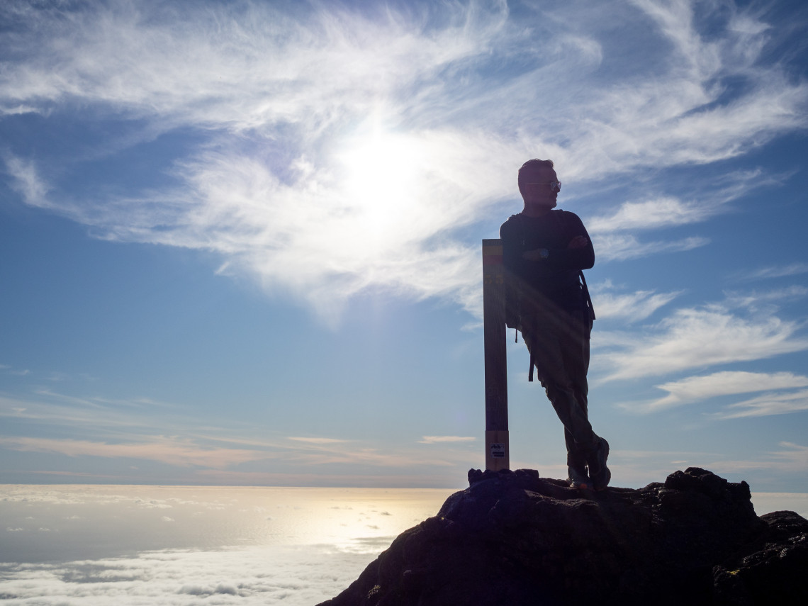 Hiker standing beside a trail marker near the summit of Mount Pico, silhouetted against bright clouds and the Atlantic horizon.