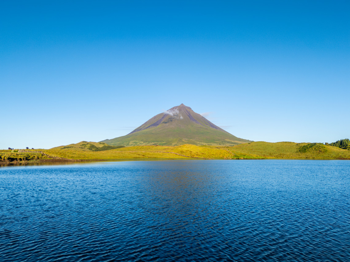 Lagoa do Capit&atilde;o stretches across the foreground with Mount Pico rising above the green highland landscape under a clear blue sky.