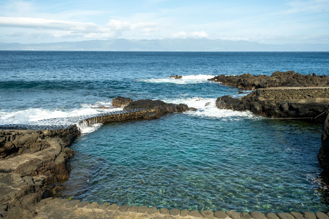 Clear seawater fills a sheltered natural swimming pool bordered by black volcanic rock and stone walls on the coast of Pico Island.