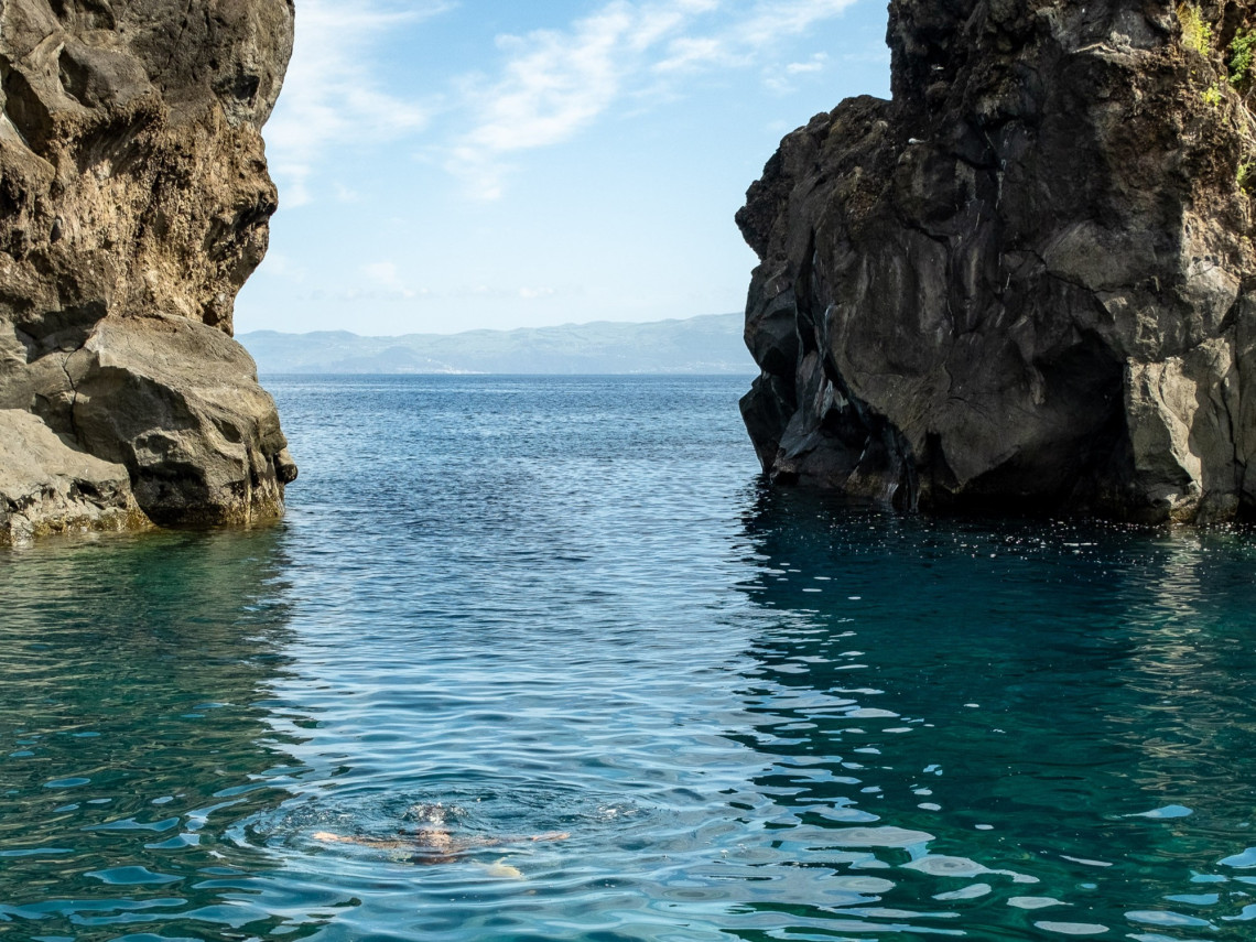 Clear Atlantic water between steep black volcanic rock formations at Arcos do Cachorro in Bandeiras, with a swimmer visible in the sheltered channel.