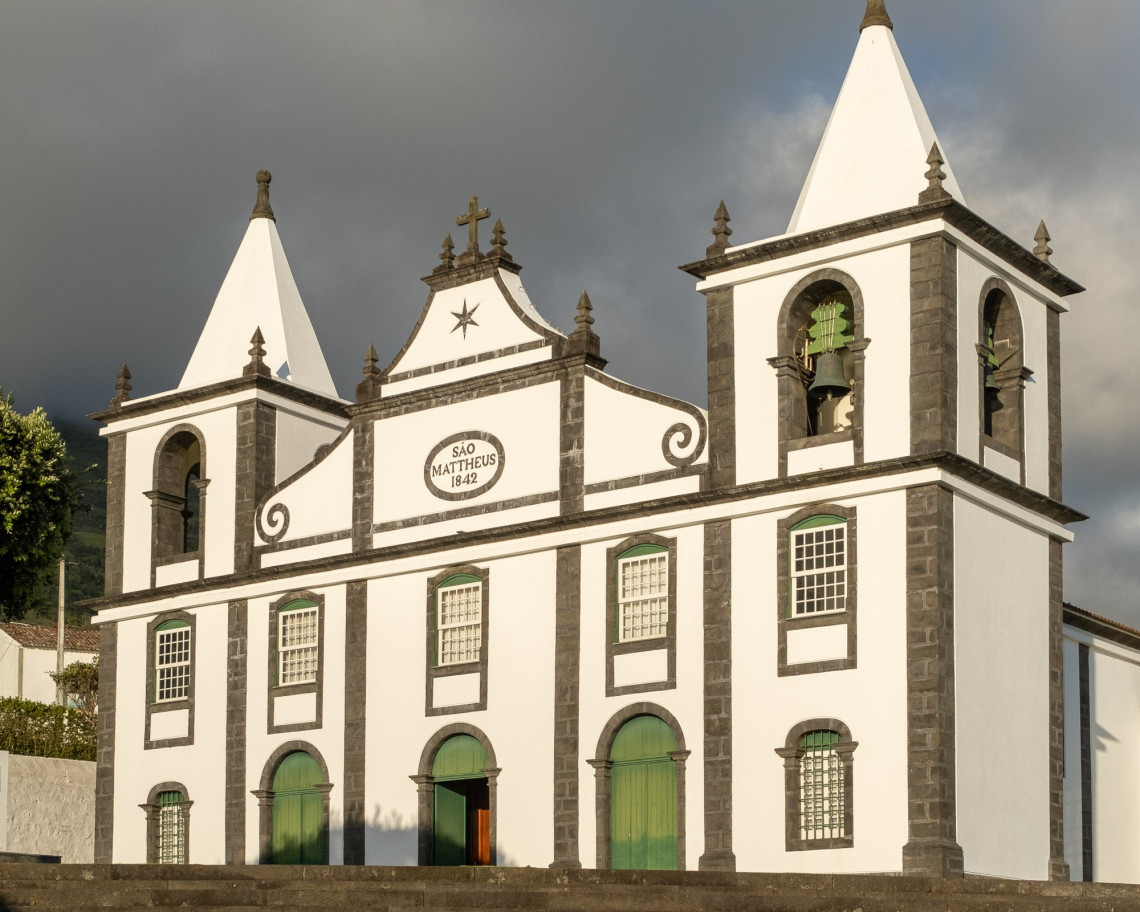 White fa&ccedil;ade of Igreja de S&atilde;o Mateus on Pico Island, framed by dark volcanic stone and twin bell towers with green doors and windows.