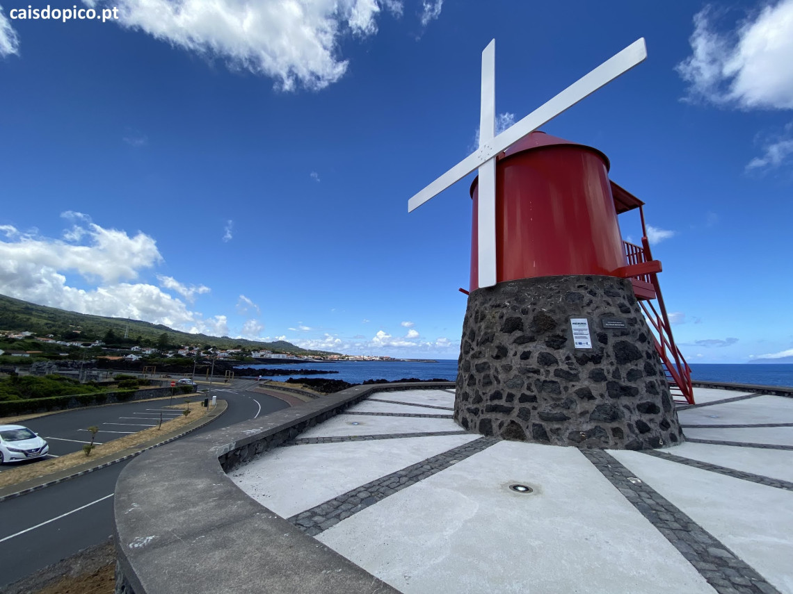 Red windmill with white sails standing on a black volcanic stone base beside Avenida do Mar in S&atilde;o Roque do Pico, overlooking the Atlantic coast.
