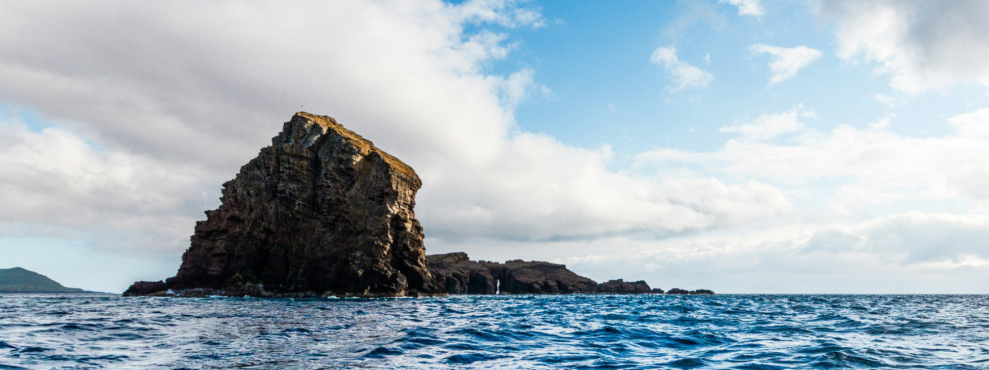Volcanic rock formation of the Madalena Islets rising from the Atlantic Ocean off the western coast of Pico Island, with the surrounding coastline visible in the distance.