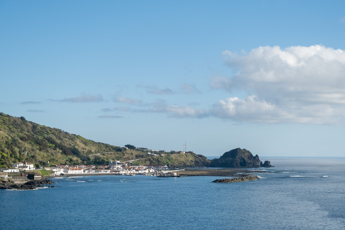 Coastal settlement on Pico Island overlooking a sheltered Atlantic bay, with whitewashed houses, green hills and dark volcanic rock formations along the shoreline.
