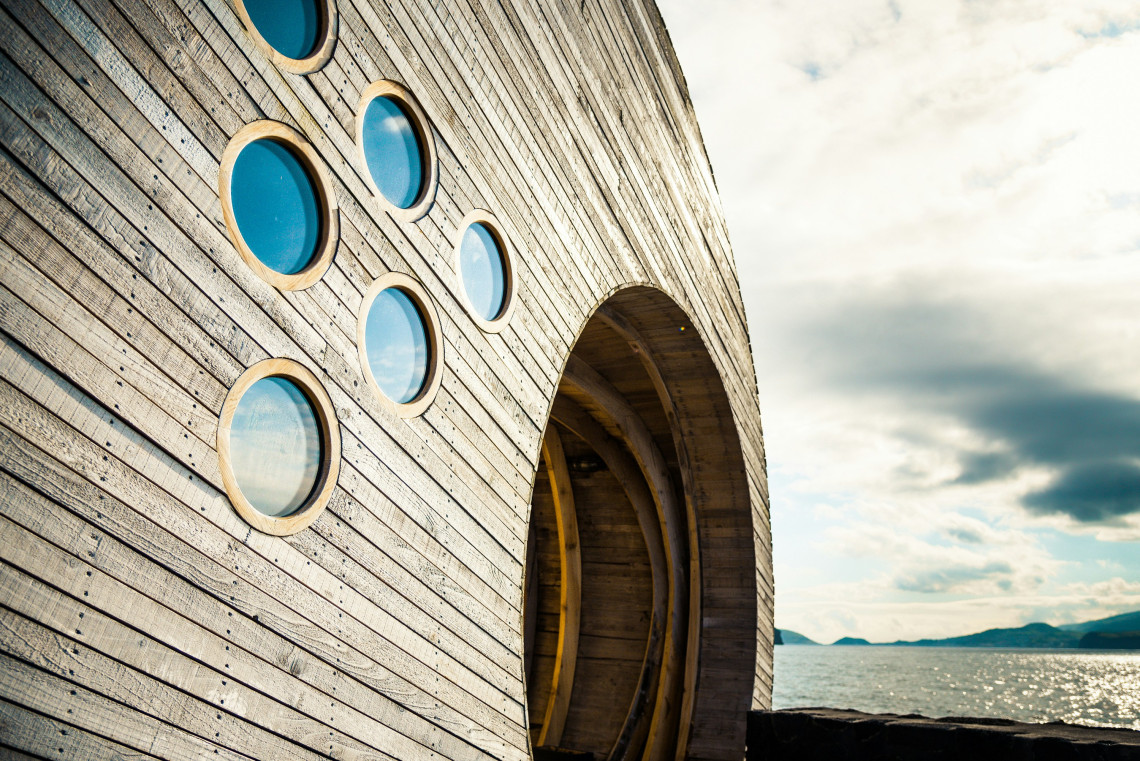Curved wooden exterior of Cella Bar in Madalena, featuring circular windows and a large arched entrance beside the Atlantic Ocean.