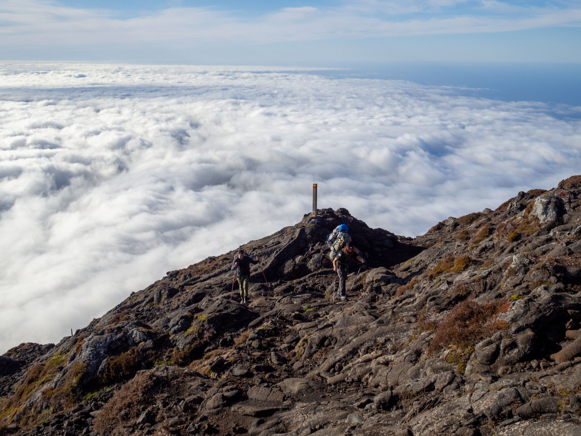 Hikers crossing the rocky volcanic slopes of Mount Pico above a dense layer of clouds during the ascent of Portugal&rsquo;s highest mountain.