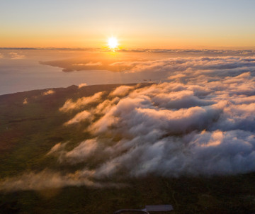 Sunrise over Mount Pico and the surrounding volcanic landscape, with warm light illuminating a dense layer of clouds below the mountain slopes.