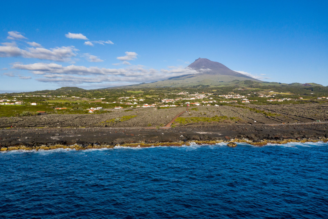View from the Atlantic toward Pico Island, with Mount Pico rising behind green settlements and an extensive black volcanic coastline.