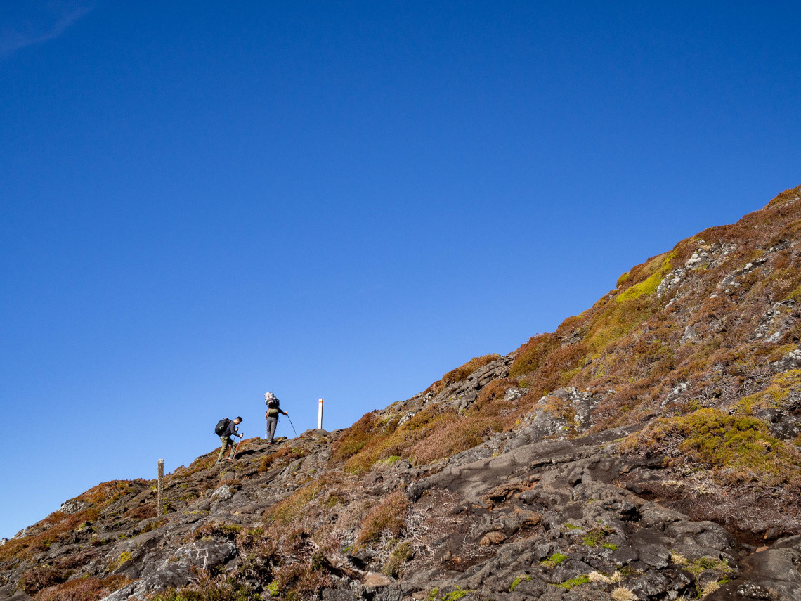 Hikers ascending the steep rocky trail of Mount Pico beneath a clear blue sky, surrounded by volcanic terrain and low mountain vegetation.