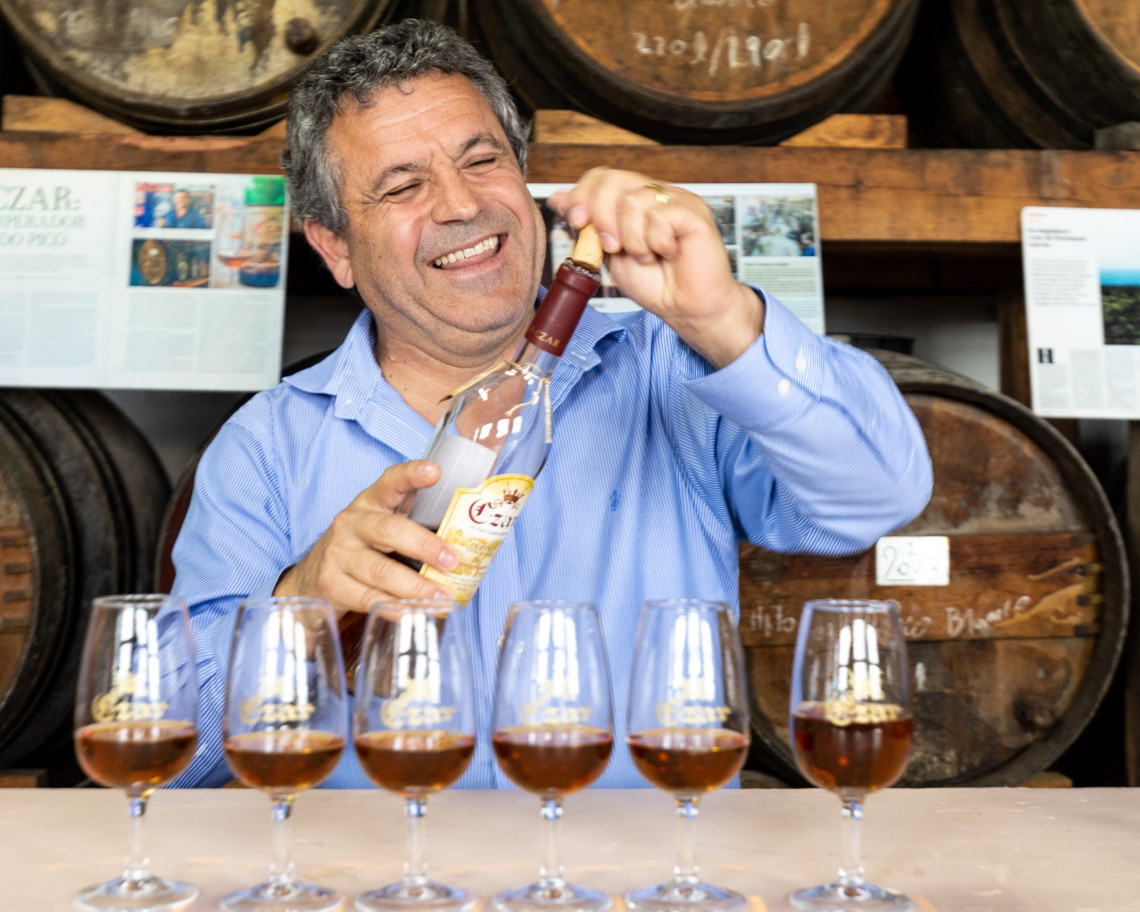 Wine tasting at Czar Winery on Pico Island, with glasses of amber-colored wine arranged in front of wooden barrels as a bottle is opened.