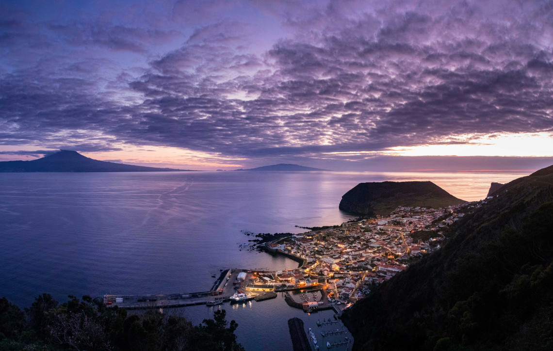 Elevated evening view over Horta and its harbor on Faial Island, with the Atlantic channel and the silhouette of Mount Pico visible in the distance.