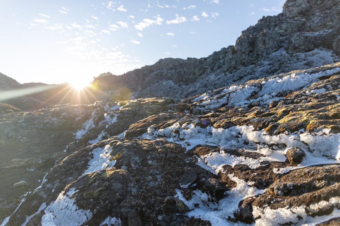 Snow and ice covering sections of the dark volcanic rock near the summit of Mount Pico as sunlight rises over the rugged mountain terrain.