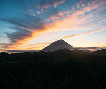 Mount Pico silhouetted against an orange and blue sunset sky, rising above the dark volcanic landscape of Pico Island in the Azores.