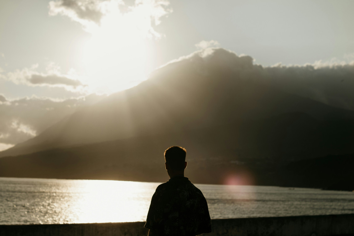 Silhouetted traveler looking toward Mount Pico as sunlight breaks through clouds above the mountain and reflects across the water.