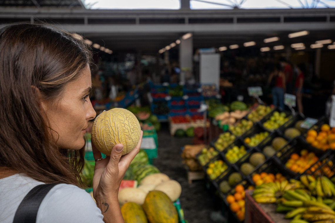 Visitor smelling fresh fruit at Mercado da Gra&ccedil;a in Ponta Delgada, surrounded by stalls selling locally grown produce from S&atilde;o Miguel Island.