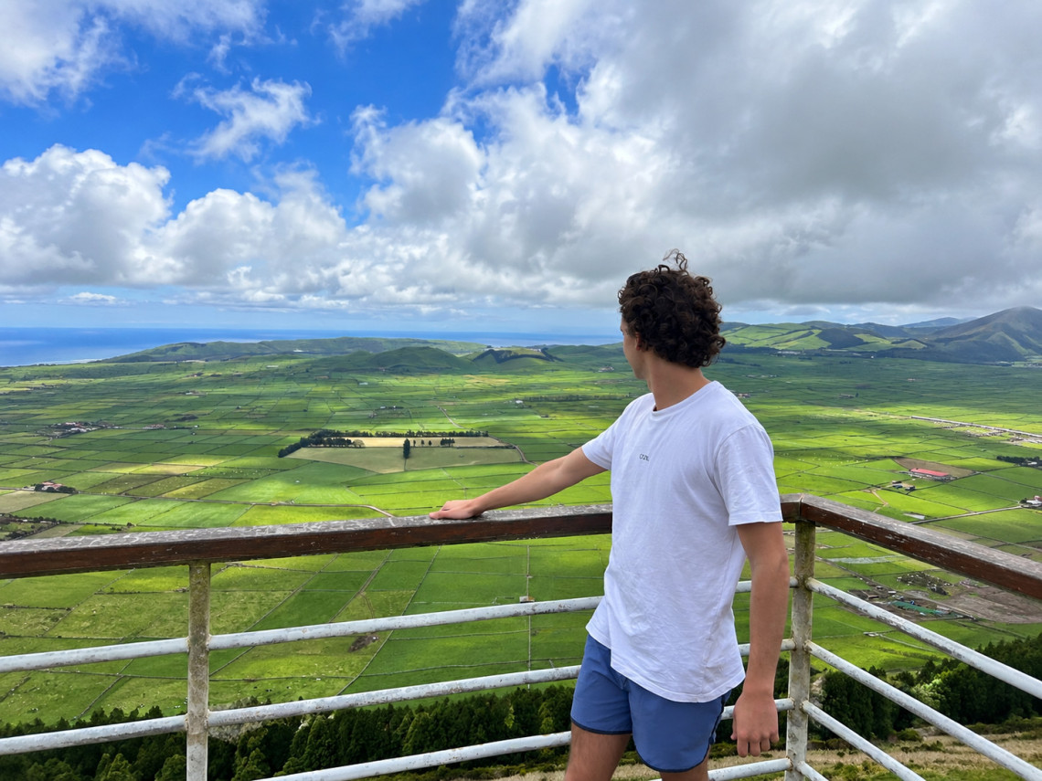 Visitor looking across the green patchwork fields from Serra do Cume viewpoint on Terceira Island, with volcanic hills and the Atlantic Ocean visible in the distance.