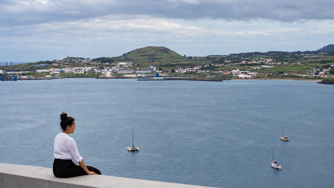 Woman sitting above Praia da Vit&oacute;ria Bay on Terceira Island, overlooking sailboats on the calm Atlantic water and the green coastal landscape beyond.