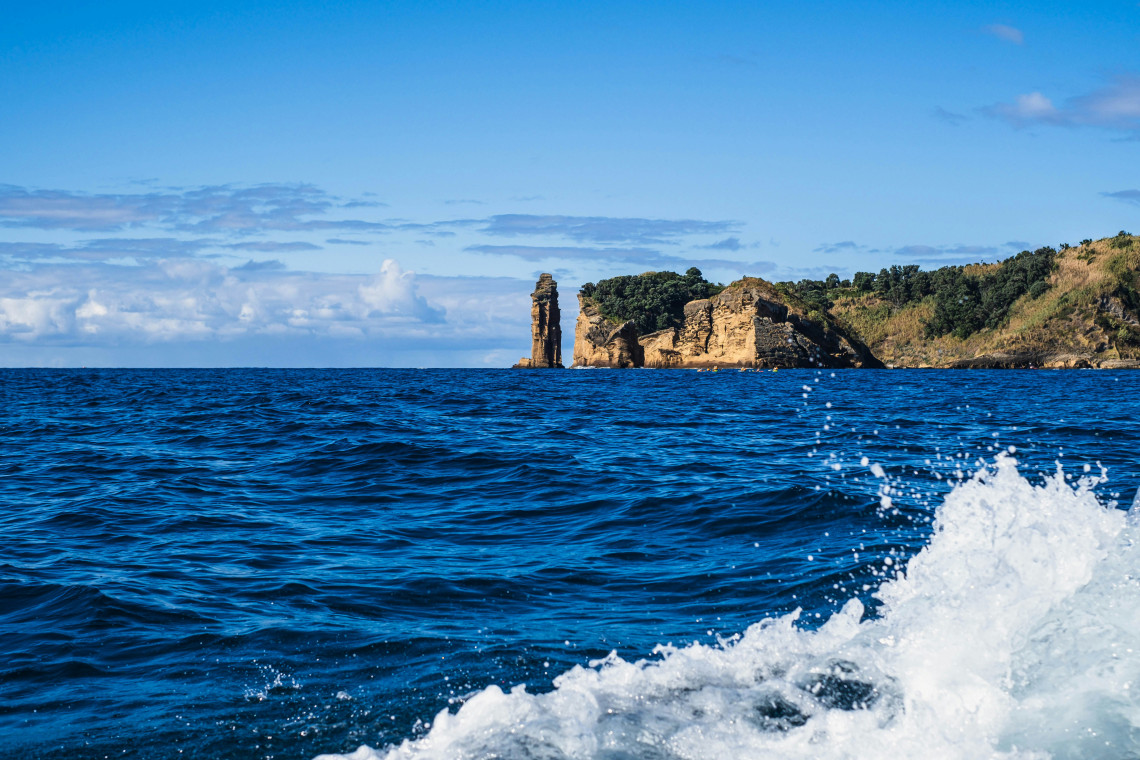 View from a moving boat of a dramatic volcanic sea stack and rugged golden cliffs rising from the deep blue Atlantic Ocean, Azores, Portugal