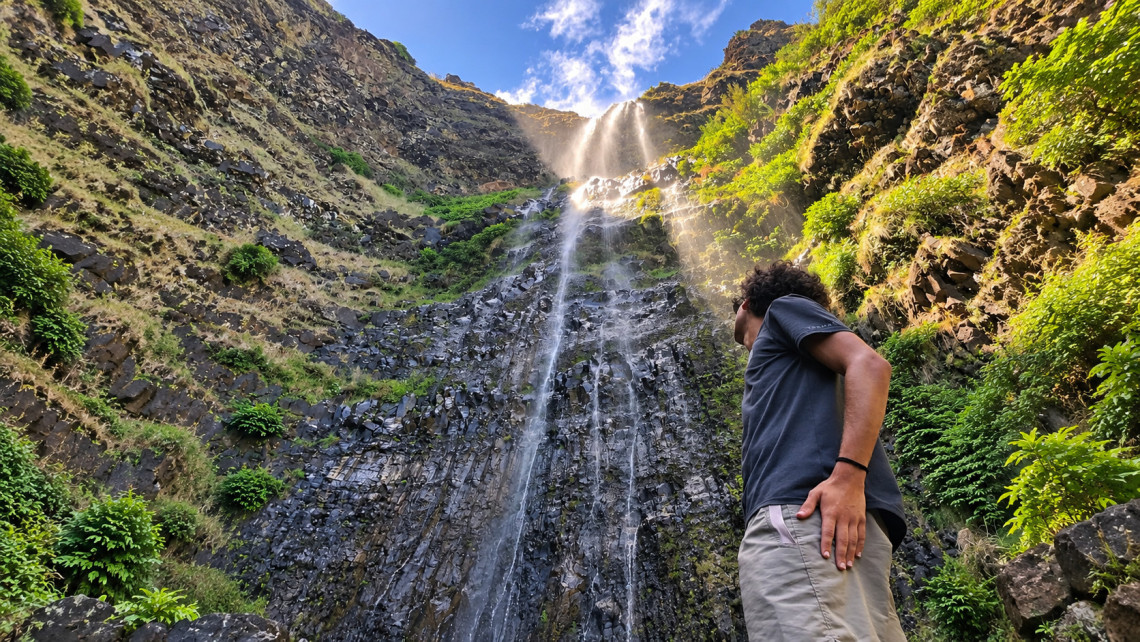 A visitor looks up at Cascata do Aveiro in Maia, where water descends a tall volcanic rock face surrounded by green vegetation on Santa Maria Island.