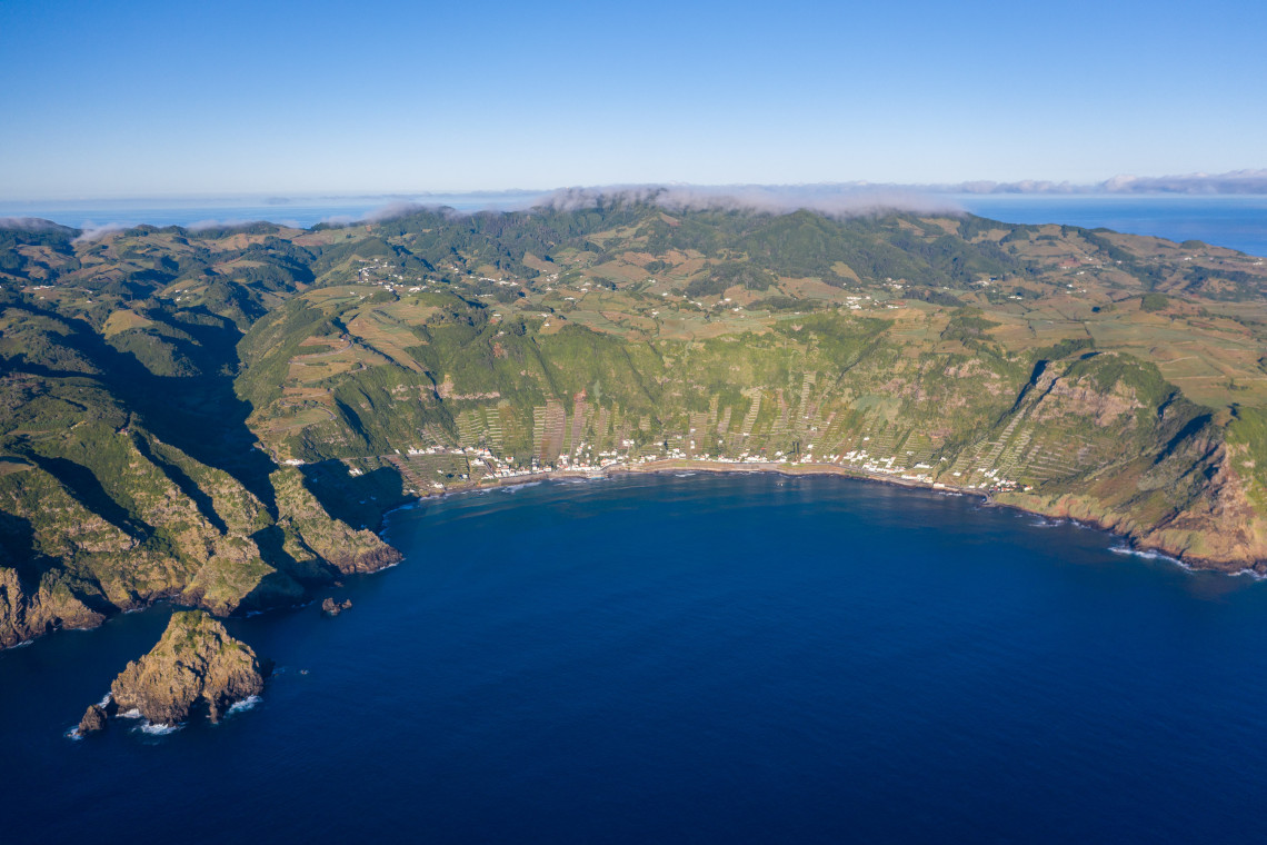 Wide aerial view of S&atilde;o Louren&ccedil;o Bay on Santa Maria Island, showing the curved Atlantic coastline, white coastal houses, steep green slopes and traditional terraced vineyards surrounding the bay.
