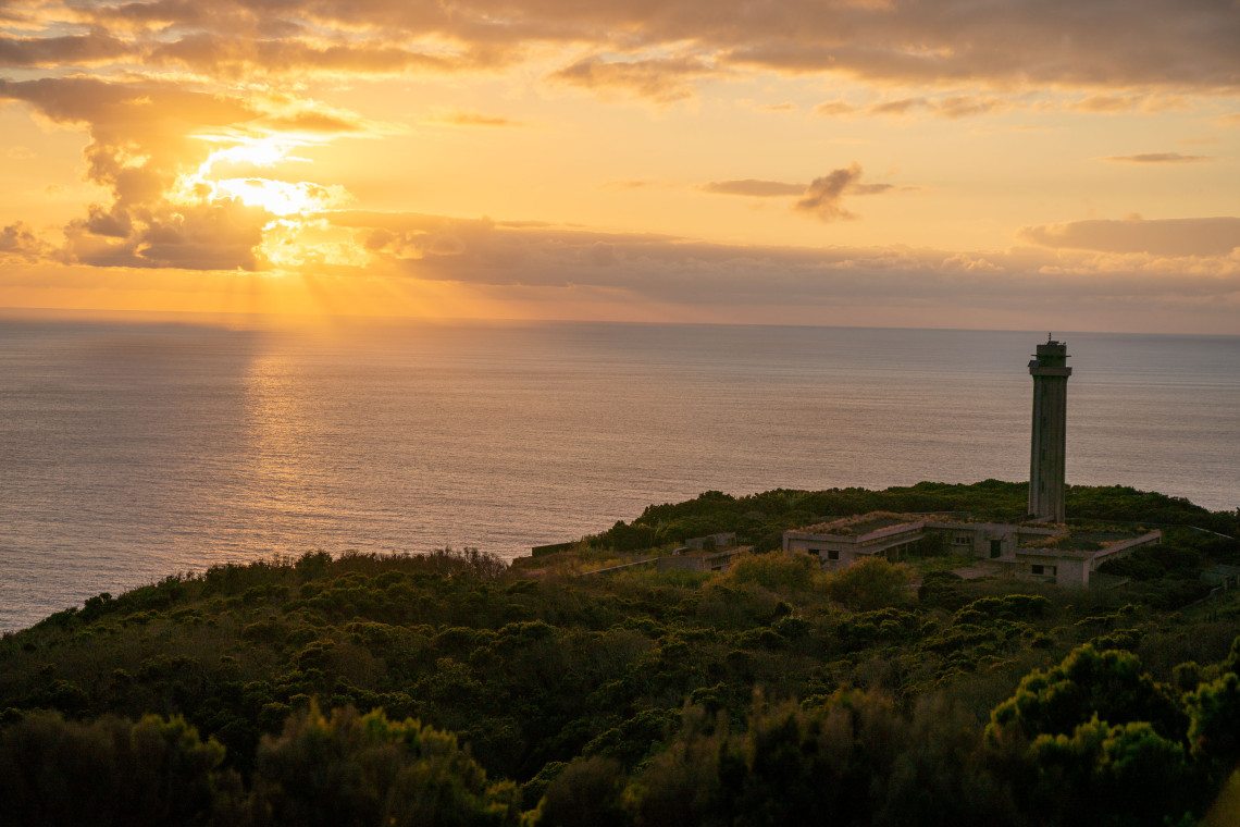 Rosais Lighthouse standing above the Atlantic coast of S&atilde;o Jorge Island, surrounded by low vegetation beneath a warm sunset sky.
