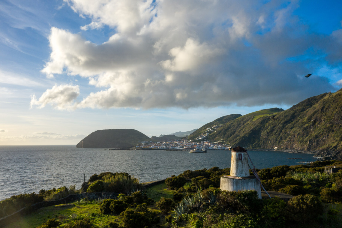 Traditional windmill overlooking the coastal village of Urzelina on S&atilde;o Jorge Island, with green hills and the Atlantic Ocean beyond.