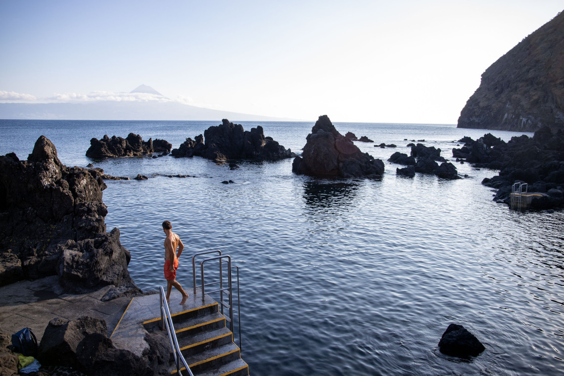 Swimmer entering the volcanic natural pools of Pregui&ccedil;a Natural Pools in Velas, S&atilde;o Jorge, with dark basalt rocks and Mount Pico visible across the Atlantic.