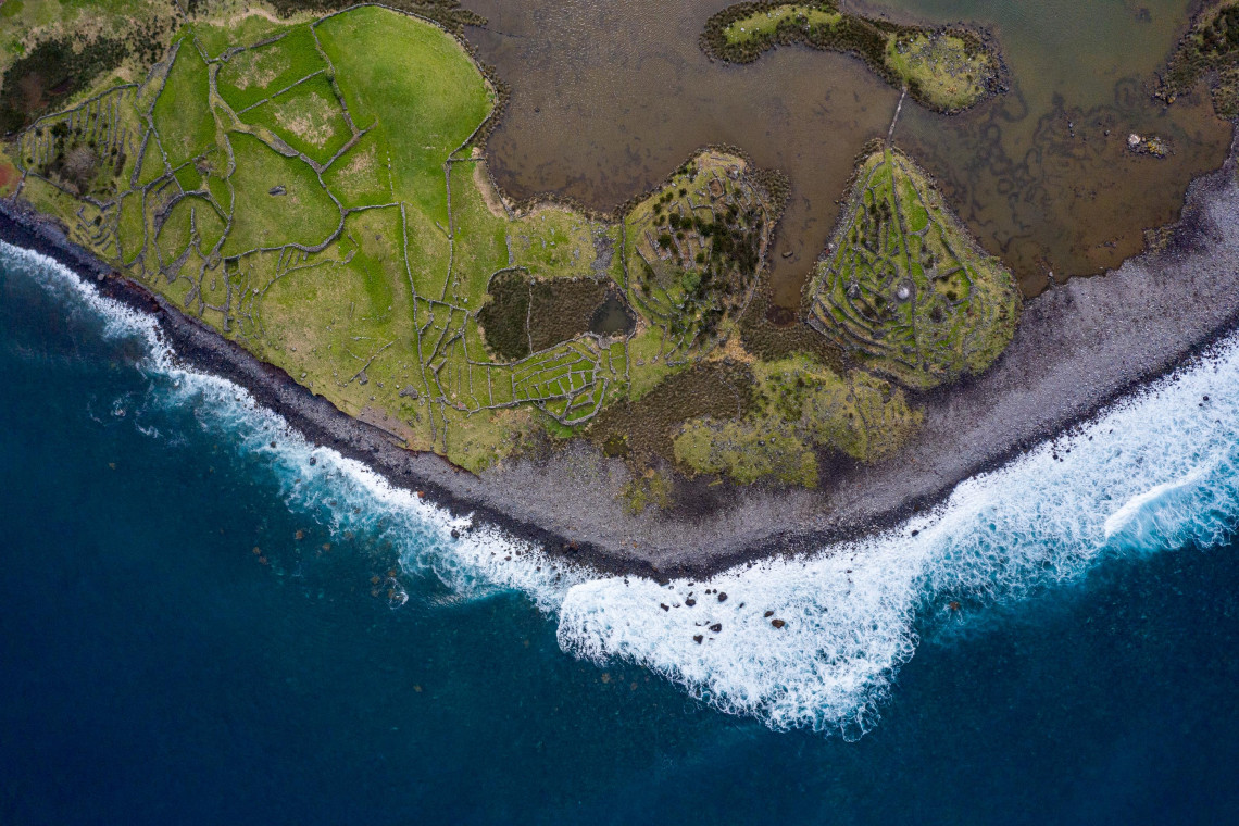 Top-down aerial view of Faj&atilde; da Caldeira de Santo Cristo on S&atilde;o Jorge Island, showing its coastal lagoon, green agricultural plots and dark volcanic shoreline beside the Atlantic Ocean.