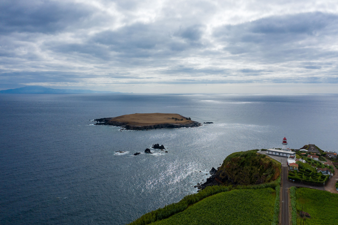 View toward Ilh&eacute;u do Topo off the eastern coast of S&atilde;o Jorge Island, with the low volcanic islet surrounded by the Atlantic Ocean and Ponta do Topo Lighthouse visible on the nearby headland.