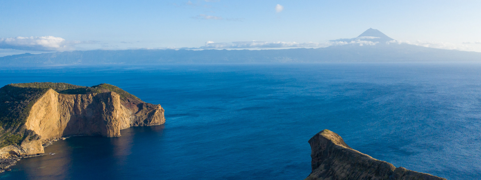 View over the volcanic Morro de Velas on São Jorge Island, surrounded by the Atlantic Ocean with Mount Pico rising across the channel in the distance.