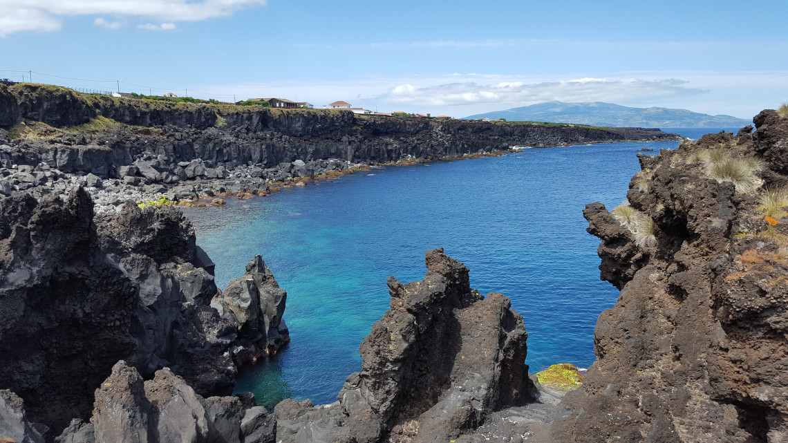 Clear Atlantic water surrounded by rugged black lava formations along the coast of Pico Island, with green slopes visible across the bay.