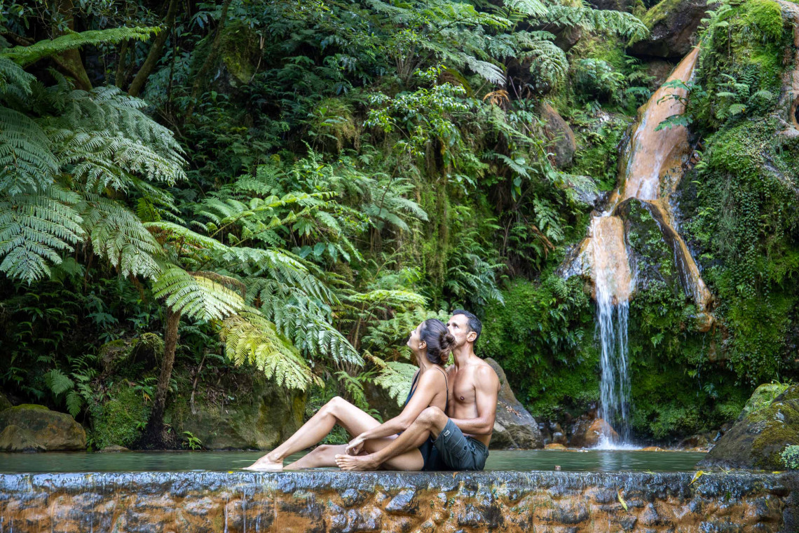 Couple relaxing beside the warm waterfall at Caldeira Velha, surrounded by giant ferns and dense subtropical forest on S&atilde;o Miguel Island.