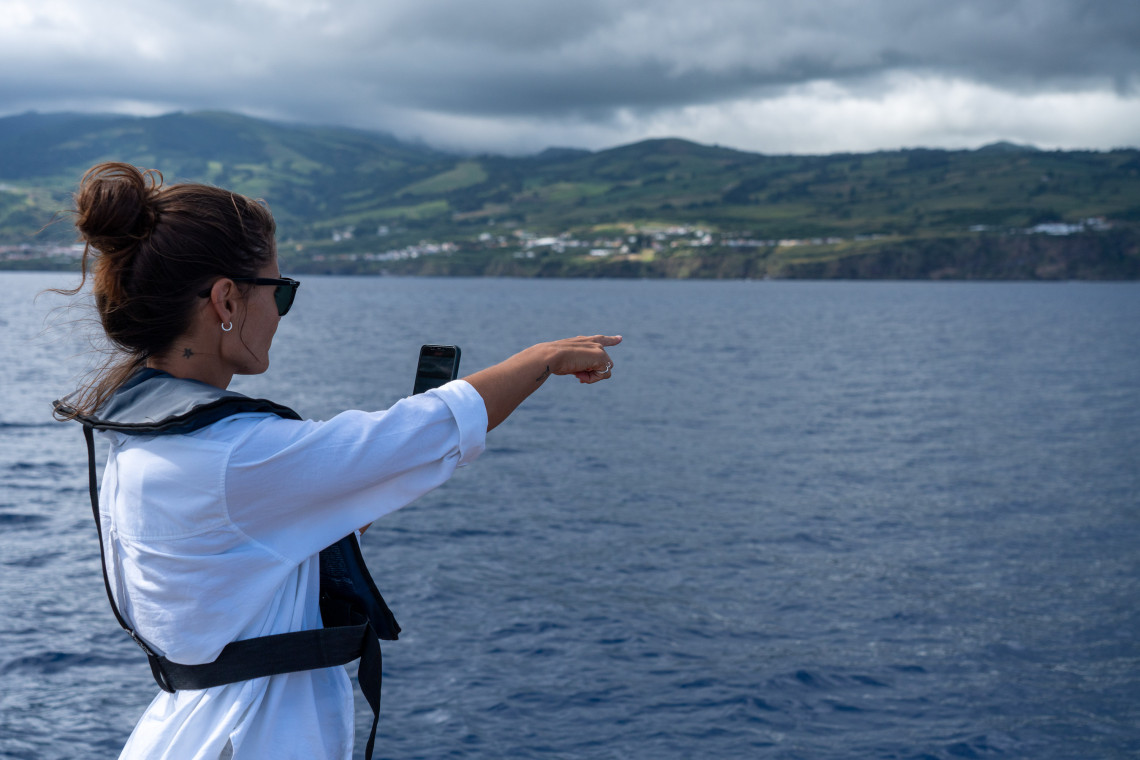 Visitor pointing toward the ocean during a whale-watching boat tour off S&atilde;o Miguel Island, with the island&rsquo;s green coastline in the background.