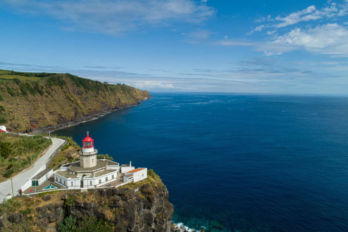 Aerial view of Farol do Arnel perched above the rocky coast of Nordeste, with its red lantern overlooking the deep blue Atlantic Ocean.