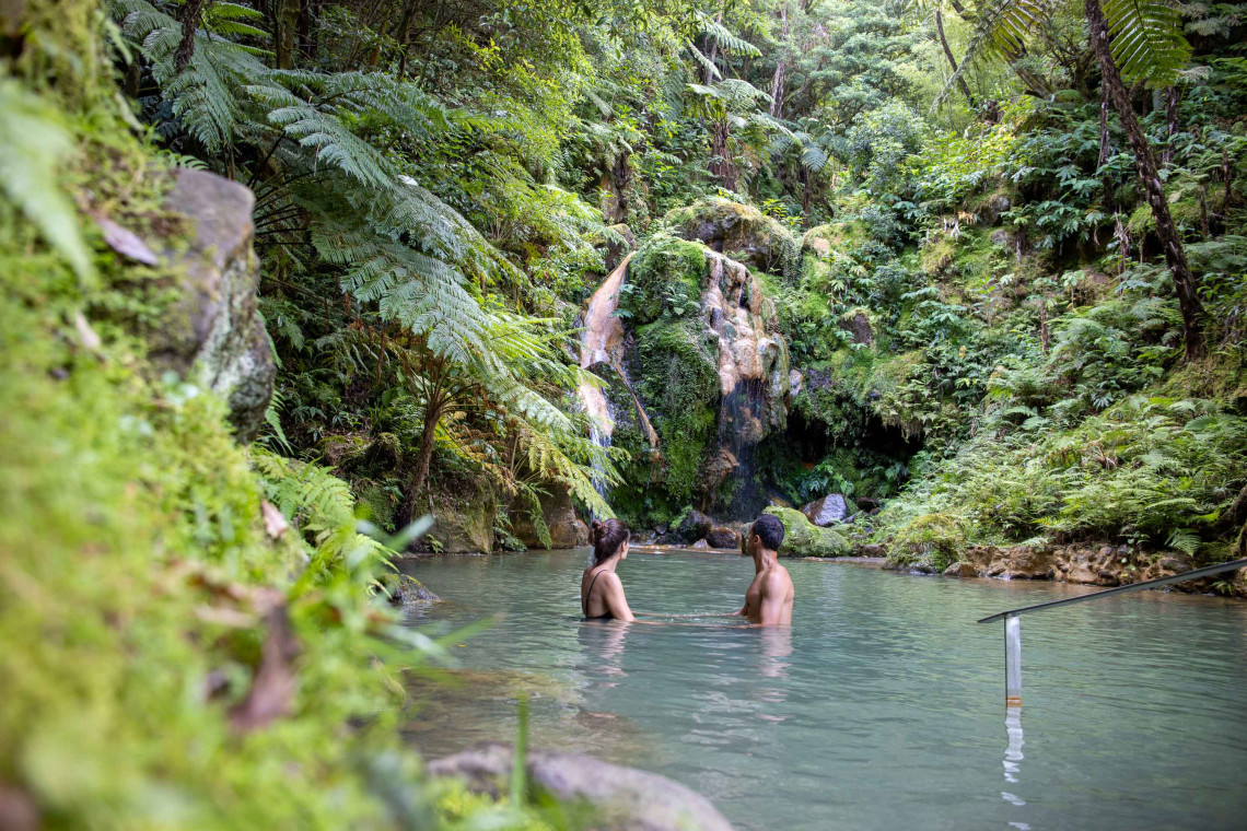 Visitors bathing in a natural thermal pool beneath a waterfall at Caldeira Velha, surrounded by giant ferns and lush forest on S&atilde;o Miguel Island.