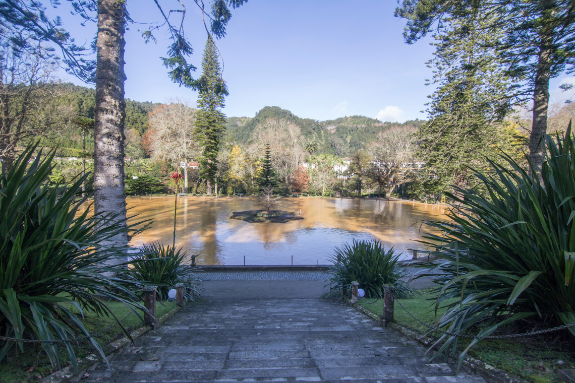 View of the large iron-rich thermal pool at Terra Nostra Park in Furnas, surrounded by mature trees and botanical gardens.