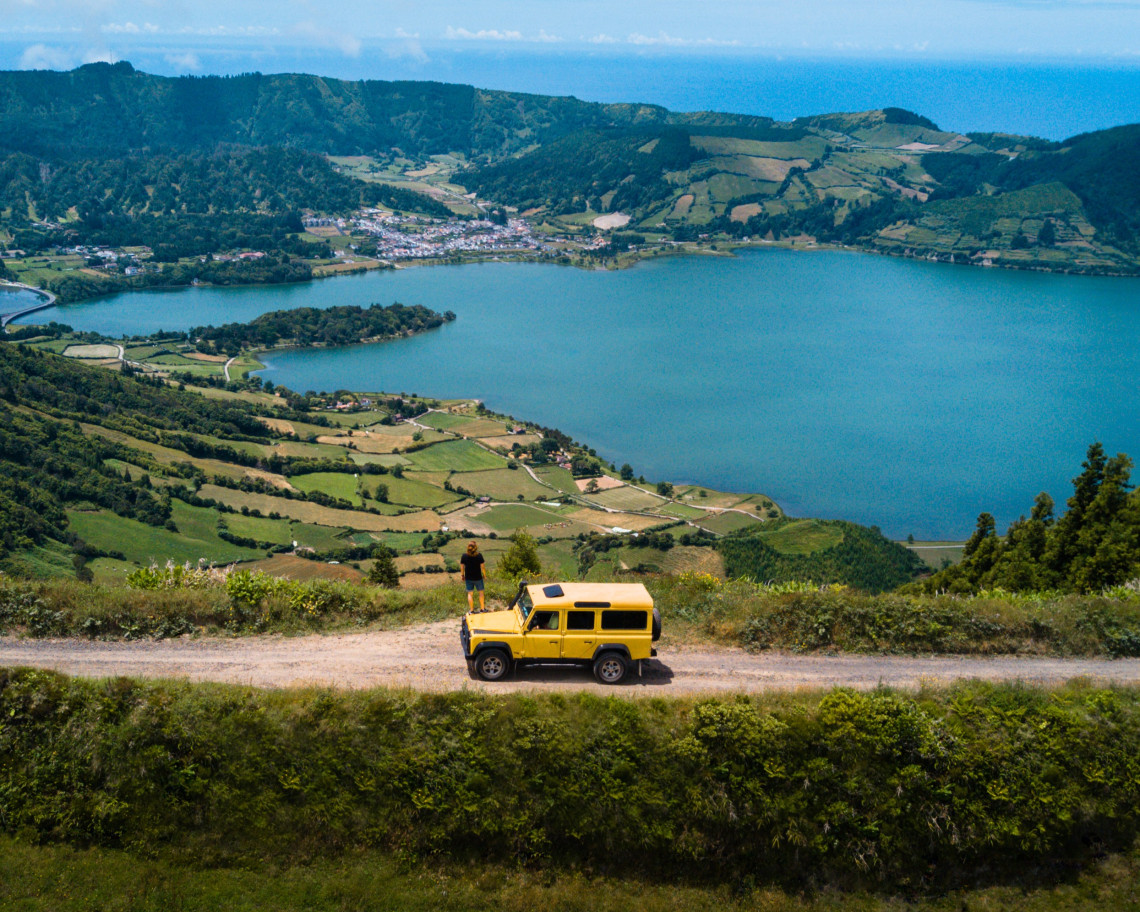 Jeep traveling along a scenic road above Lagoa das Sete Cidades, with the volcanic lake, green fields and village spread across the caldera below.