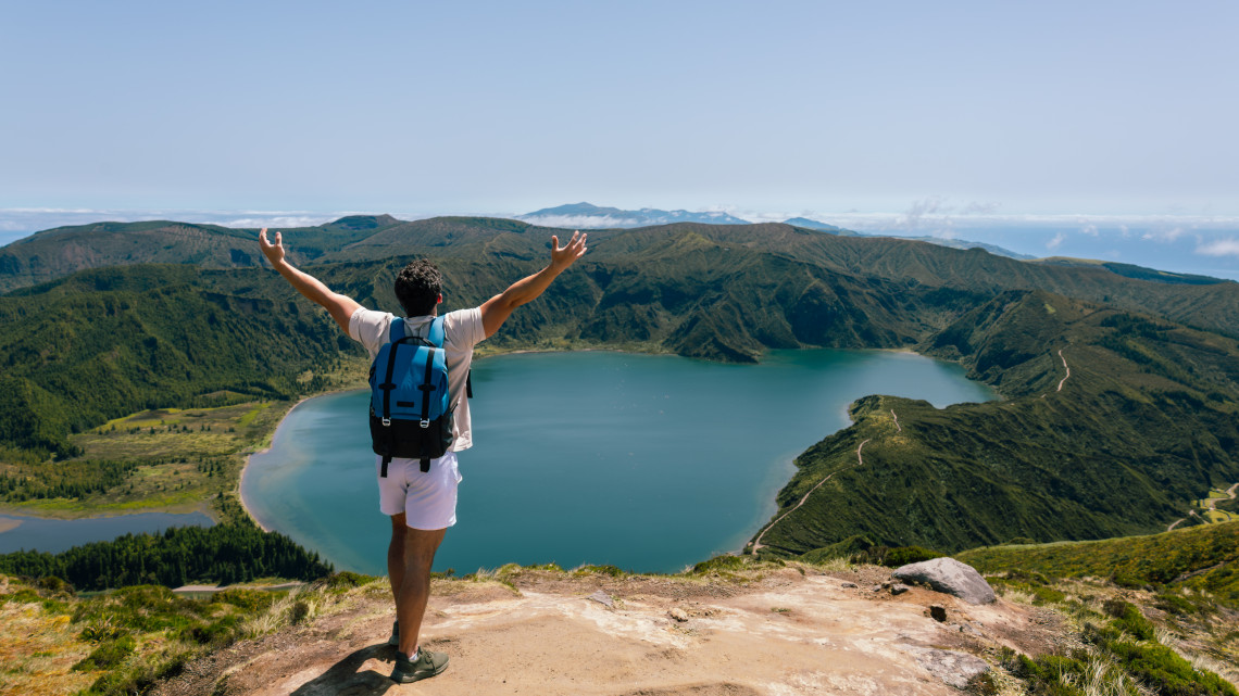 Hiker overlooking Lagoa do Fogo on S&atilde;o Miguel Island, with arms raised above the blue crater lake and surrounding volcanic mountains.