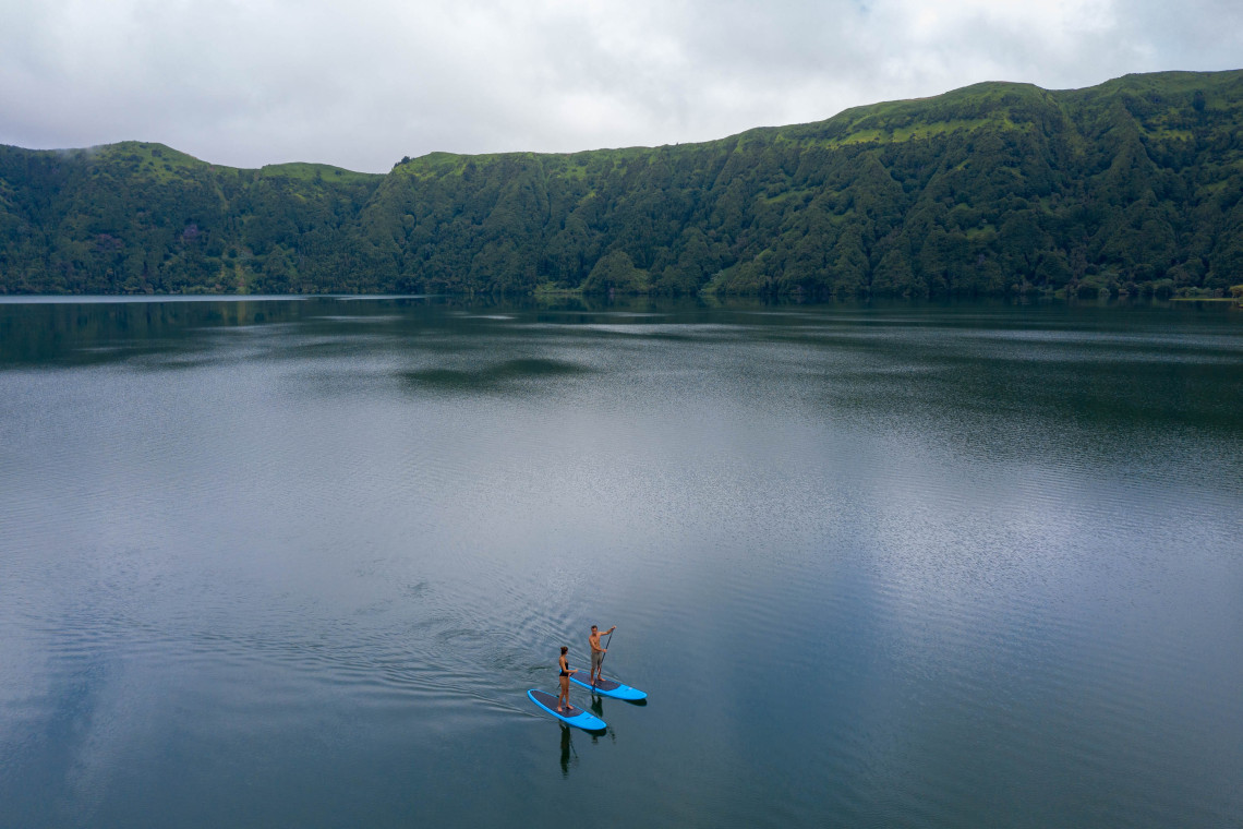 Two people stand-up paddleboarding across the calm waters of Lagoa das Sete Cidades, surrounded by the forested walls of the volcanic caldera.