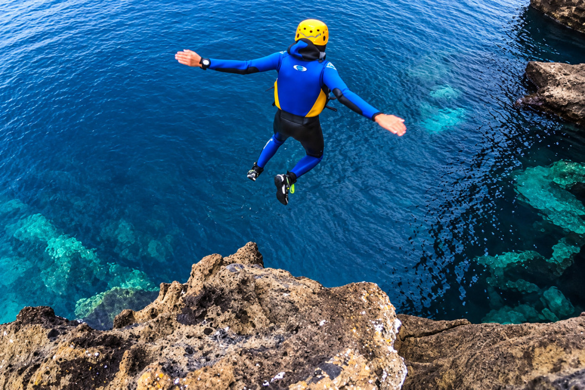 Adventurer wearing a wetsuit, helmet and flotation vest jumping from volcanic rocks into clear Atlantic waters during a coasteering experience on S&atilde;o Miguel Island.