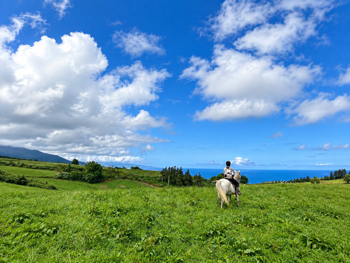 Rider exploring the green countryside of S&atilde;o Miguel Island on horseback, with rolling pastures and the Atlantic Ocean beneath a bright blue sky.