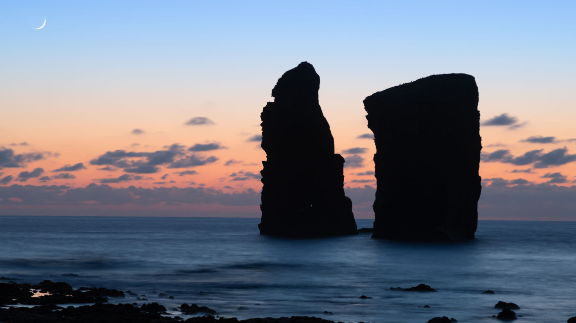Silhouetted volcanic sea stacks of Mosteiros rising from the Atlantic Ocean at sunset, beneath a crescent moon and warm evening sky.
