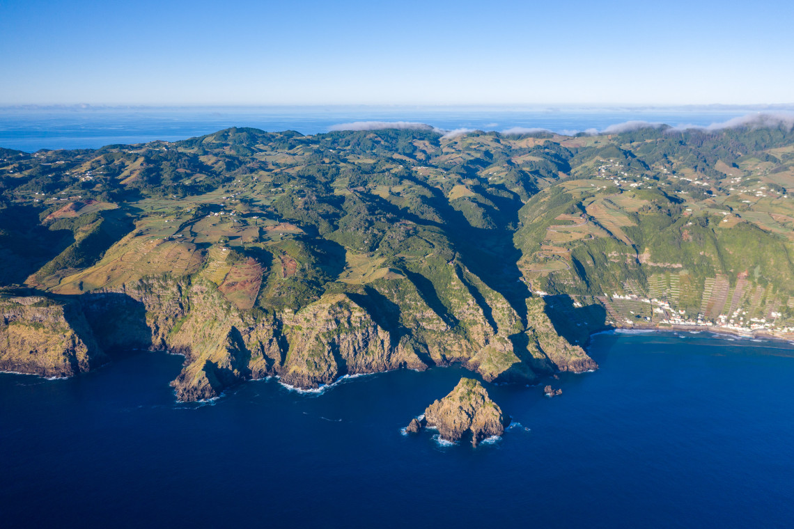 Aerial view of the rugged coastline of Santa Maria Island in the Azores, showing steep volcanic cliffs, green fields, rocky offshore formations and deep blue Atlantic waters.