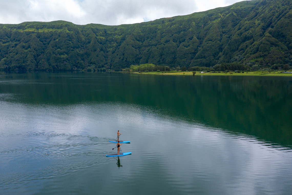 Two visitors paddleboard across the calm waters of Sete Cidades Lake on S&atilde;o Miguel Island, surrounded by the forested slopes of the volcanic caldera.