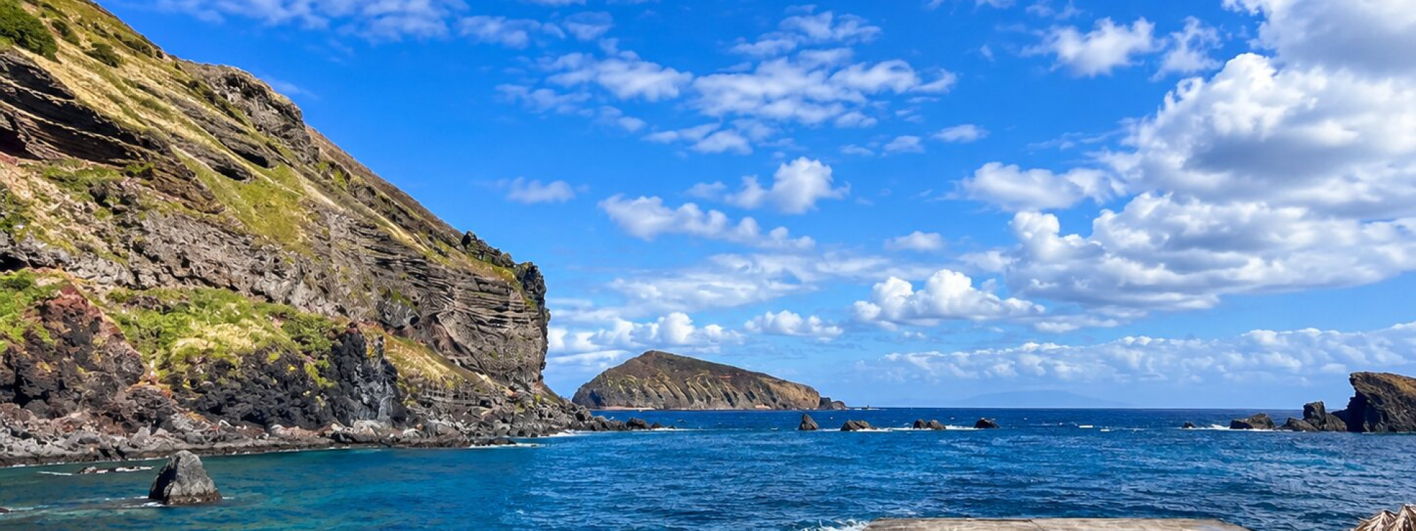 Coastal view from the Carapacho Natural Pools on Graciosa Island, with clear Atlantic water, steep layered cliffs and rocky islets extending offshore.