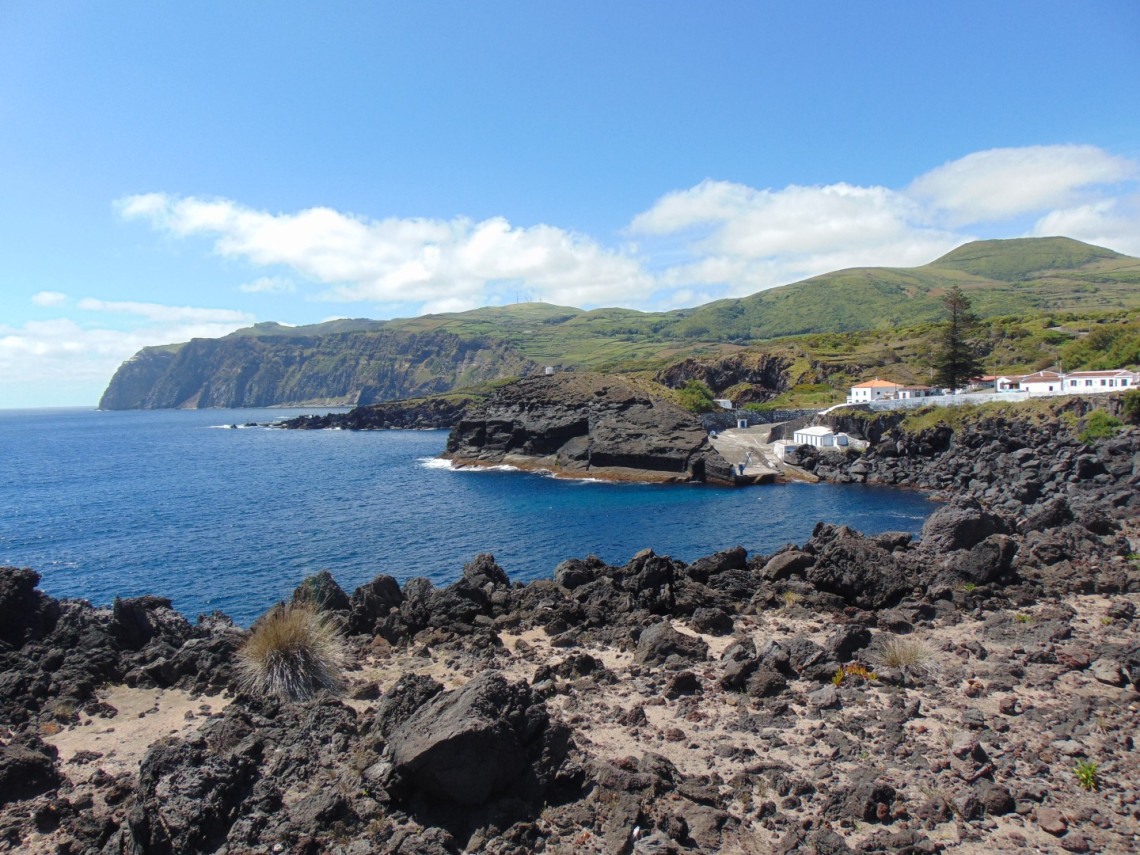 View across Porto da Folga on the southern coast of Graciosa Island, showing a small harbor sheltered by volcanic rock formations and steep green coastal cliffs. The former fishing port is also used as a local bathing area.