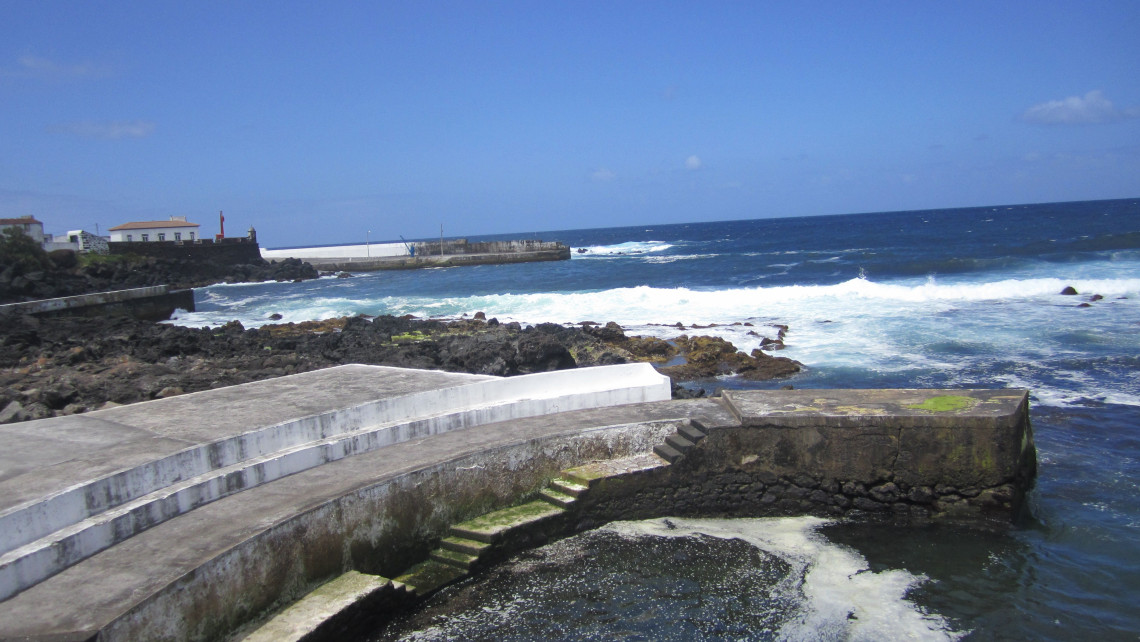 Natural swimming pools in Santa Cruz da Graciosa, with concrete walkways and steps providing access to seawater pools beside the rocky Atlantic coastline. A small harbor is visible beyond the bathing area.
